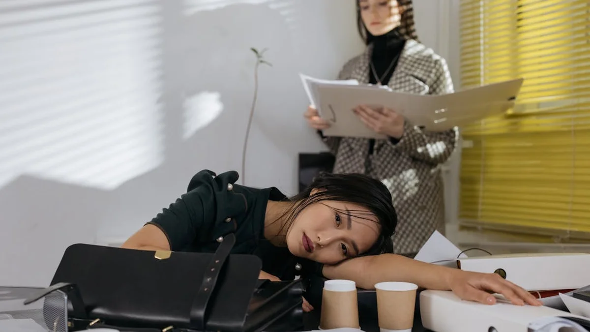 El poder de las pausas estratégicas: cómo descansar sin culp - Exhausted employee resting on desk surrounded by documents, colleague in backgro