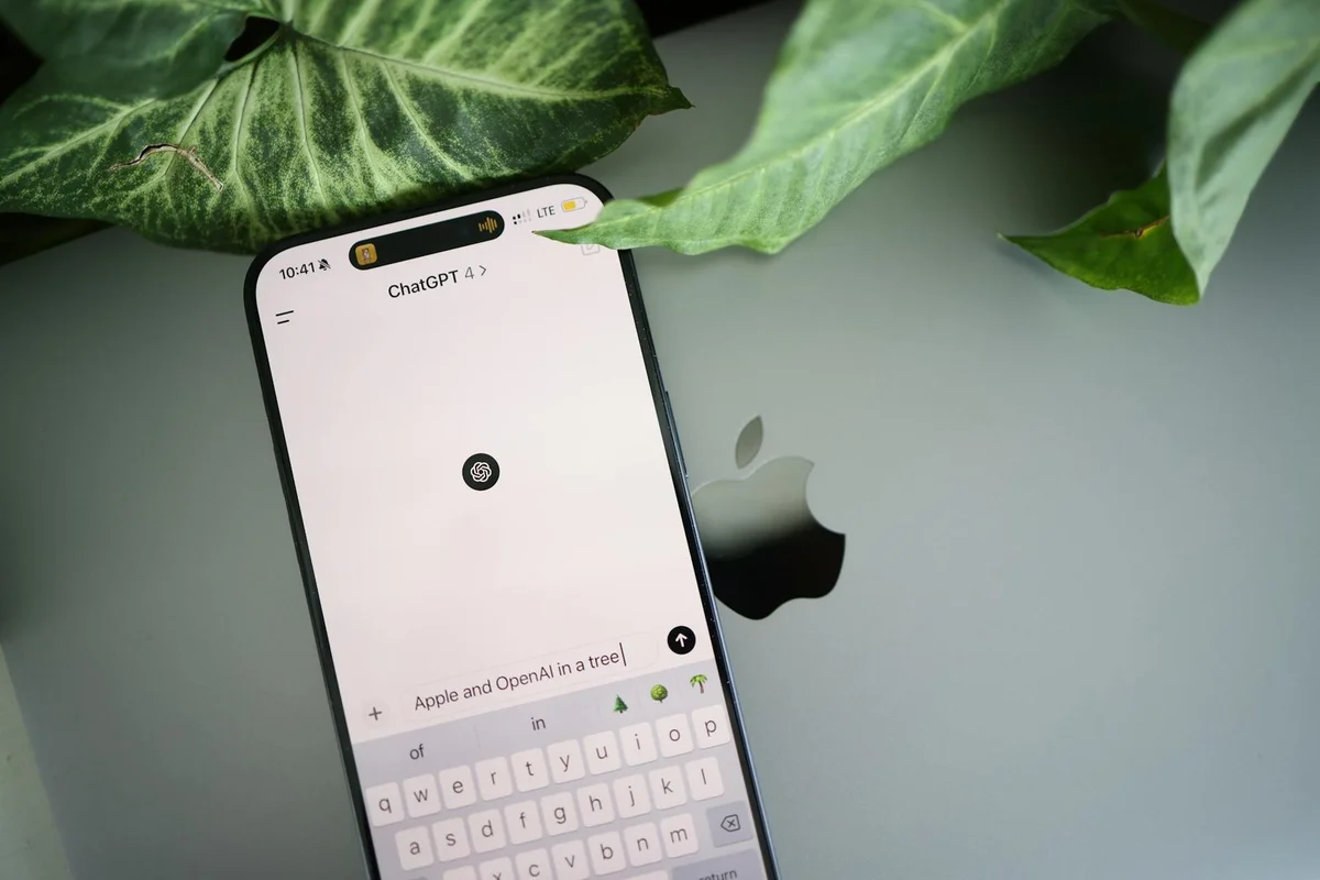 Cómo enseñar el estilo de tu negocio a la IA con few-shot: t... — A smartphone shows a ChatGPT interface placed on an Apple laptop in a leafy envi
