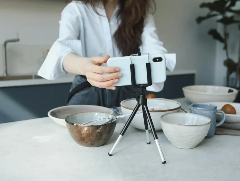 Cómo optimizar tu TikTok para búsquedas: tutorial de SEO pas... — Woman using a smartphone on a tripod for vlogging in the kitchen with bowls.