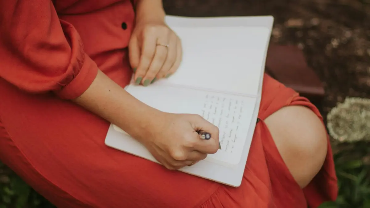 A woman in a red dress writing in a journal, outdoors over a leafy background.