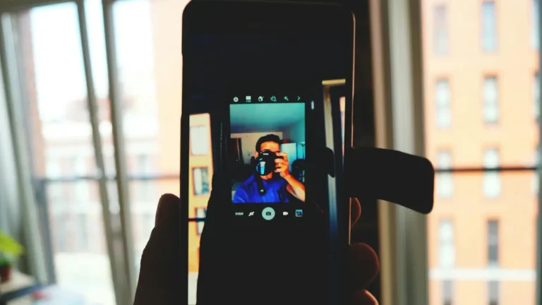 A man takes a smartphone selfie capturing his own reflection in an indoor urban setting.