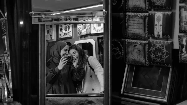 Two women smiling in a mirror, capturing a moment with a camera in an art-filled store.