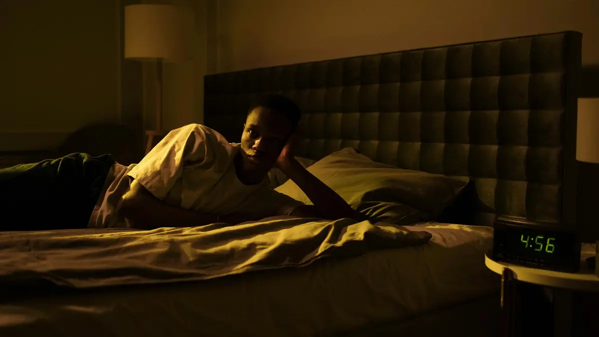 A man lies awake in bed, illuminated by early morning light from a clock reading 4:56 AM.