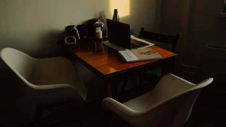 A warm, dark-toned home office with a wooden table and contemporary chairs. Includes a laptop, magaz