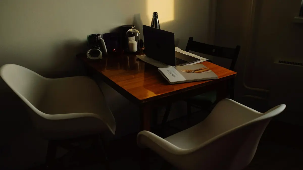 A warm, dark-toned home office with a wooden table and contemporary chairs. Includes a laptop, magaz