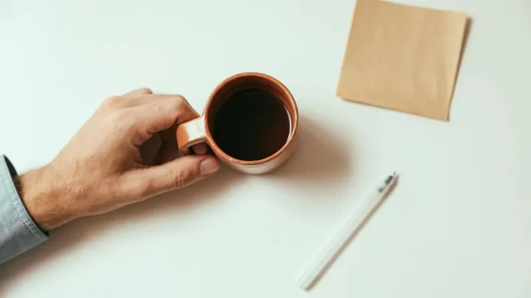 A hand holding a coffee mug on a desk with a pen and paper, creating a minimalist vibe.